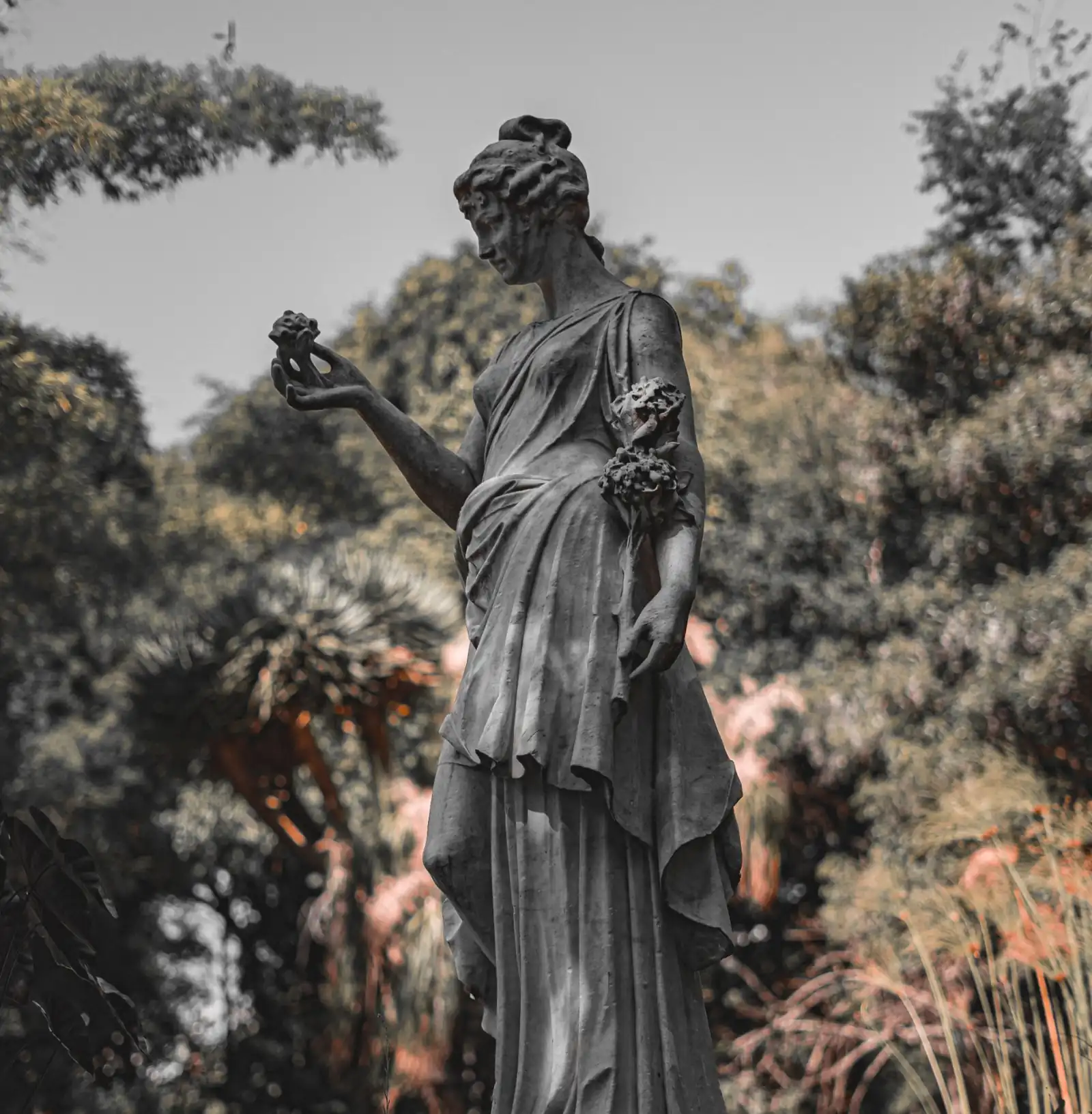 Classical stone statue of a woman holding a flower, standing on a plinth in a garden pond